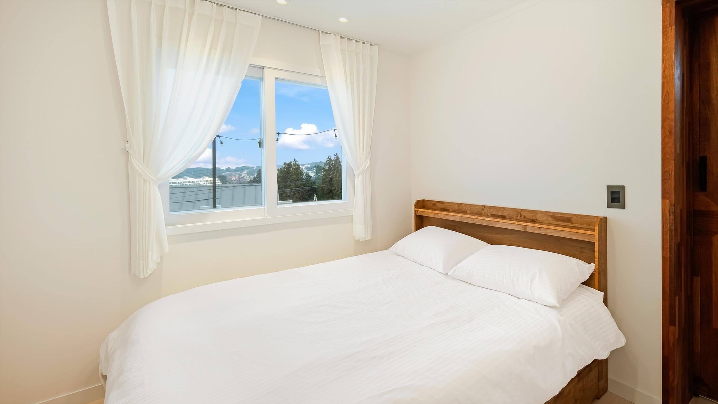 Ein Schlafzimmer mit Bett, weißen Vorhängen, einem Fenster mit Blick auf Gebäude und Bäume sowie einem hölzernen Kopfteil.