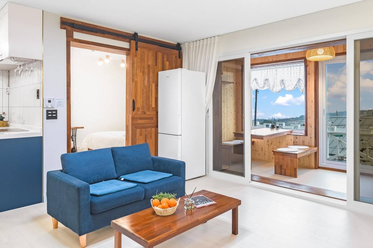 A modern living room with a blue sofa, a wooden coffee table, and a view of a balcony with a dining area.