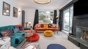 Un salon moderne avec un canapé turquoise, un fauteuil orange et un pouf jaune. On y trouve une lampe de sol noire, une petite table ronde et une grande fenêtre avec des rideaux.