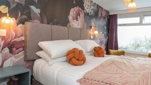 Une chambre à coucher avec un grand lit, deux lampes fixées au mur, une fresque murale à motifs floraux et une fenêtre avec des rideaux.