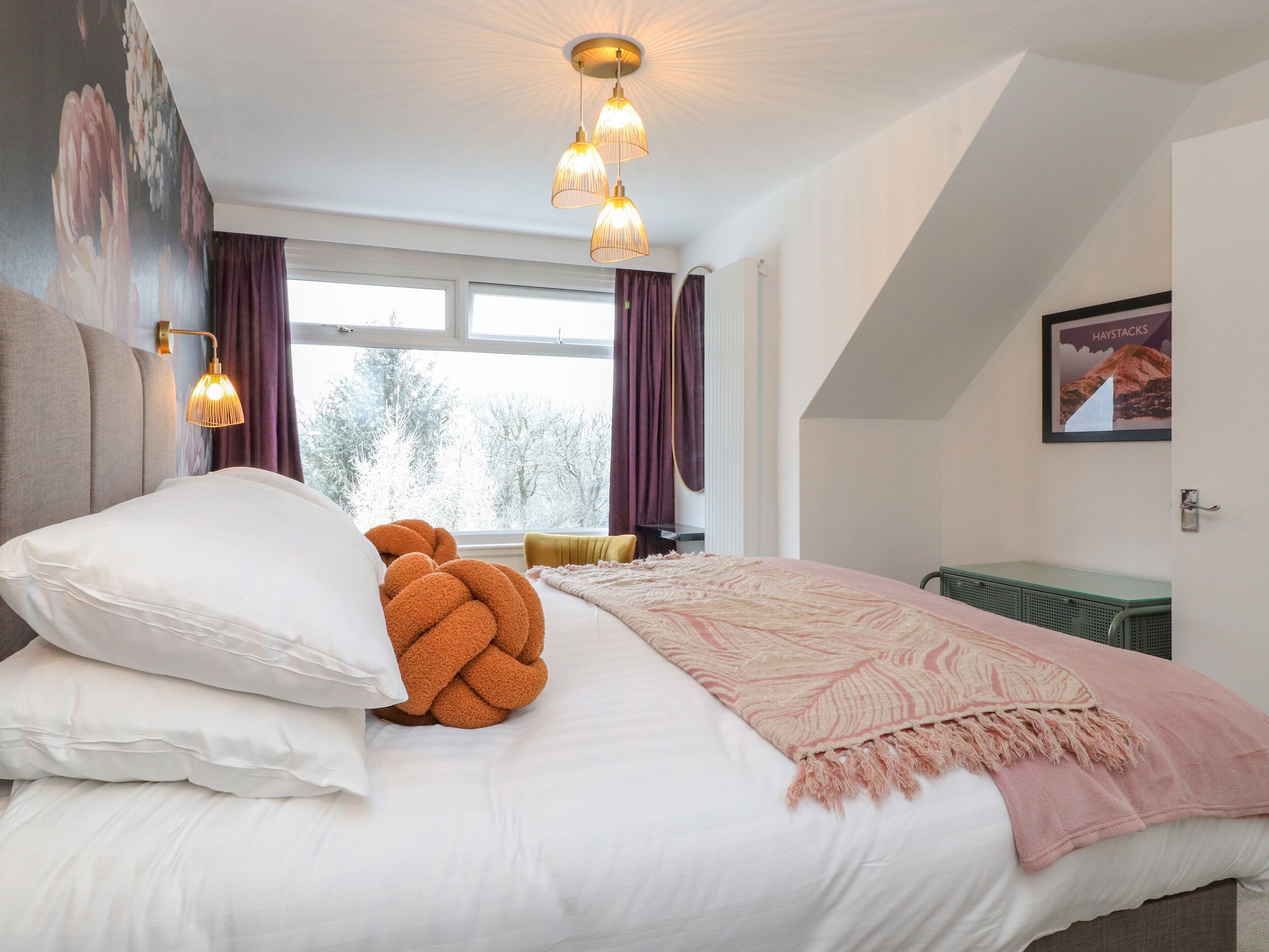 Un dormitorio con cama, almohadas, una colcha, una lámpara de pared, una ventana con cortinas y un cuadro en la pared.
