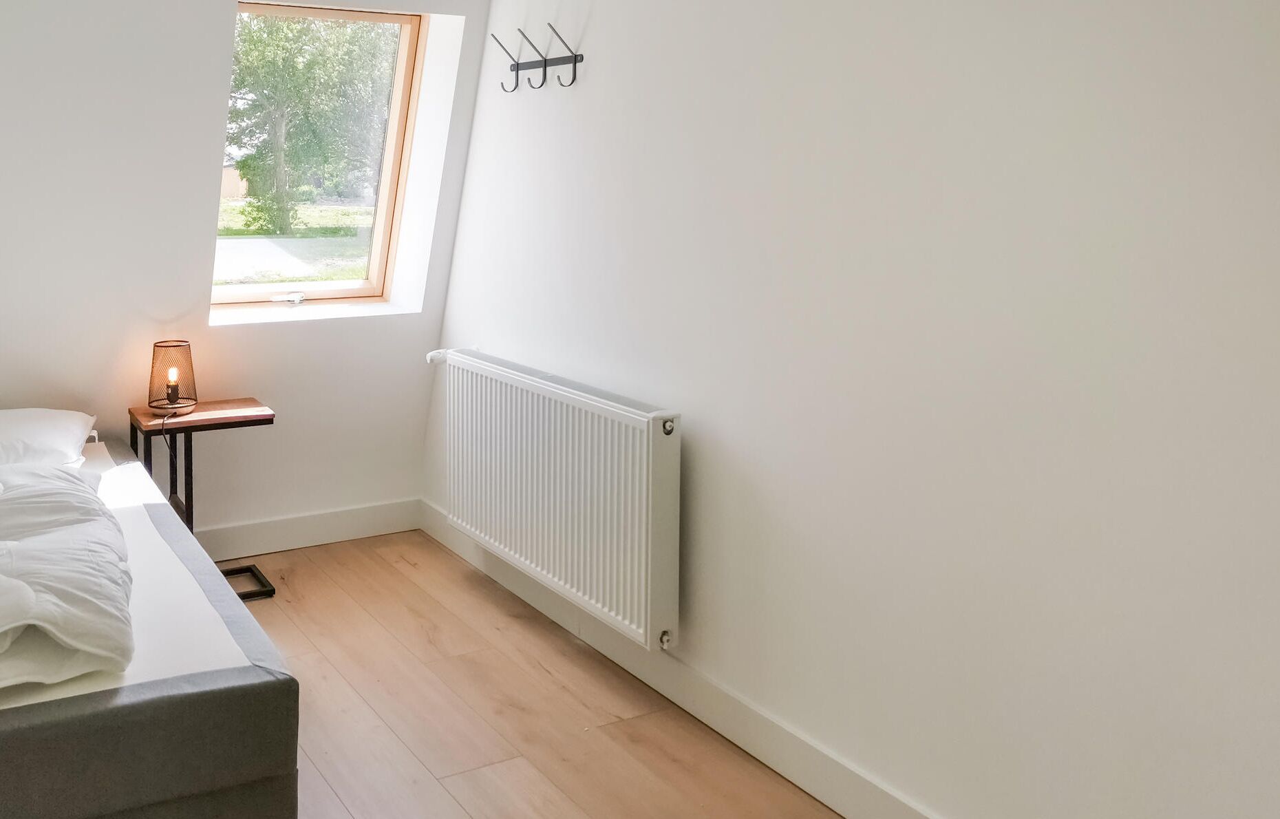 Une petite pièce lumineuse, avec un lit, une table de chevet dotée d’une lampe, une fenêtre et un radiateur.