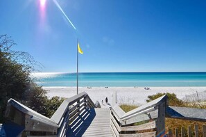 Eine Holztreppe führt zu einem Strand, bewölkter, klarer, blauer Himmel und ein Fahnenmast mit einer gelben Flagge.