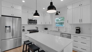 Een moderne keuken met witte kasten, roestvrijstalen apparatuur, een centraal eiland met barkrukken en hanglampen.