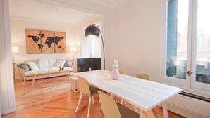 Uma sala de estar moderna com um sofá branco, uma mesa de jantar com cadeiras, uma pintura de um mapa-múndi, um abajur e uma janela com persianas.