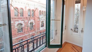 Quarto com uma janela ampla, piso de madeira e vista para um edifício vermelho com janelas em arco.