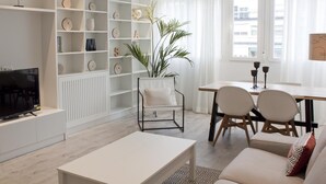 A modern living room with a white TV stand, a white coffee table, a sofa, and a dining area with a table and chairs. There is a white shelving unit with decorative items and a potted plant. - Brand new 2 bedroom apartment by RIO 2 and Matadero (Madrid)