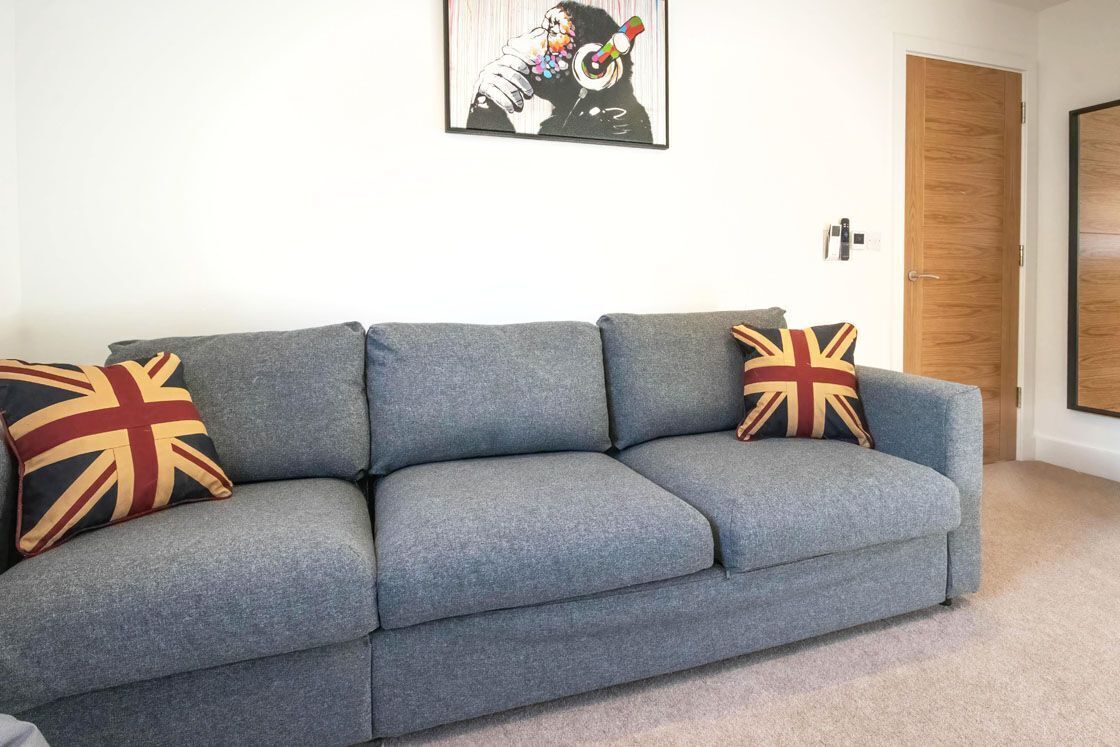 Un canapé gris agrémenté de deux coussins ornés de motifs représentant l’Union Jack, une œuvre d’art encadrée accrochée au mur et une porte en bois en arrière-plan.