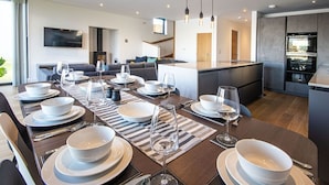 Un comedor moderno con una mesa larga dispuesta para una comida, una cocina con horno empotrado y un rincón de estar acogedor con sofá y televisor.