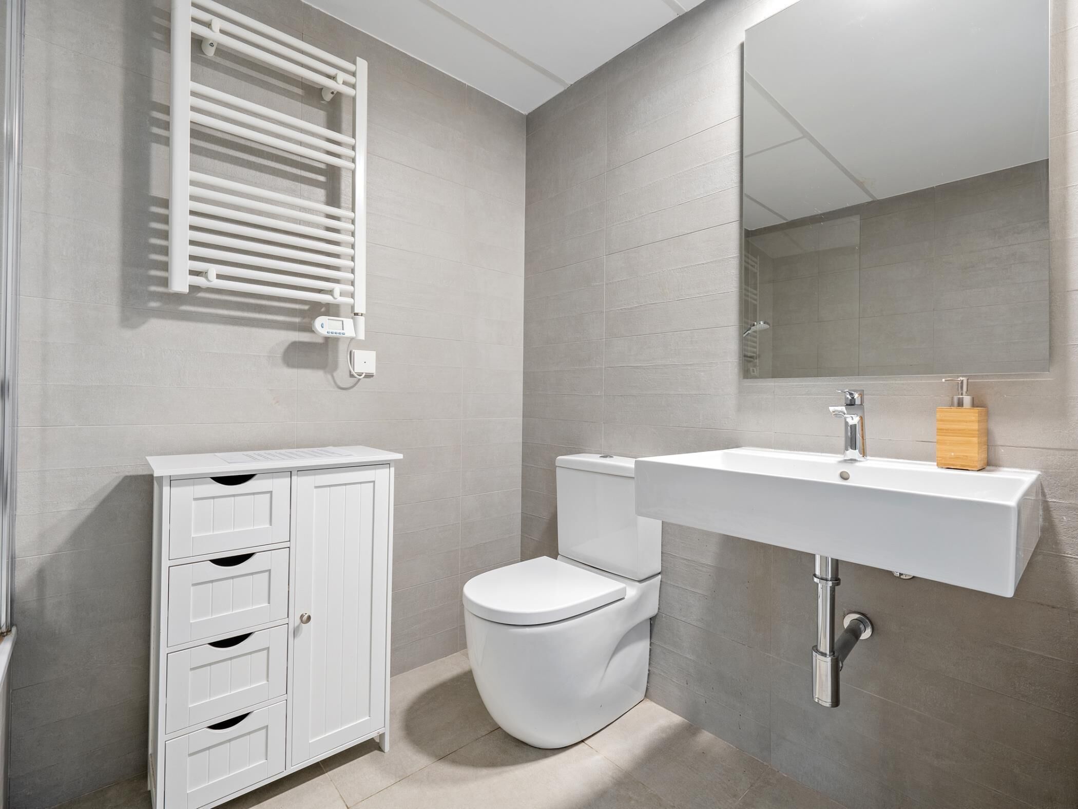 A modern bathroom with a white toilet, a white sink with a wooden soap dispenser, and a cabinet with drawers.