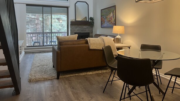 Uma sala de estar moderna com um sofá de couro marrom, uma mesa de jantar de vidro com quatro cadeiras, uma lareira e uma janela grande com vista para árvores.