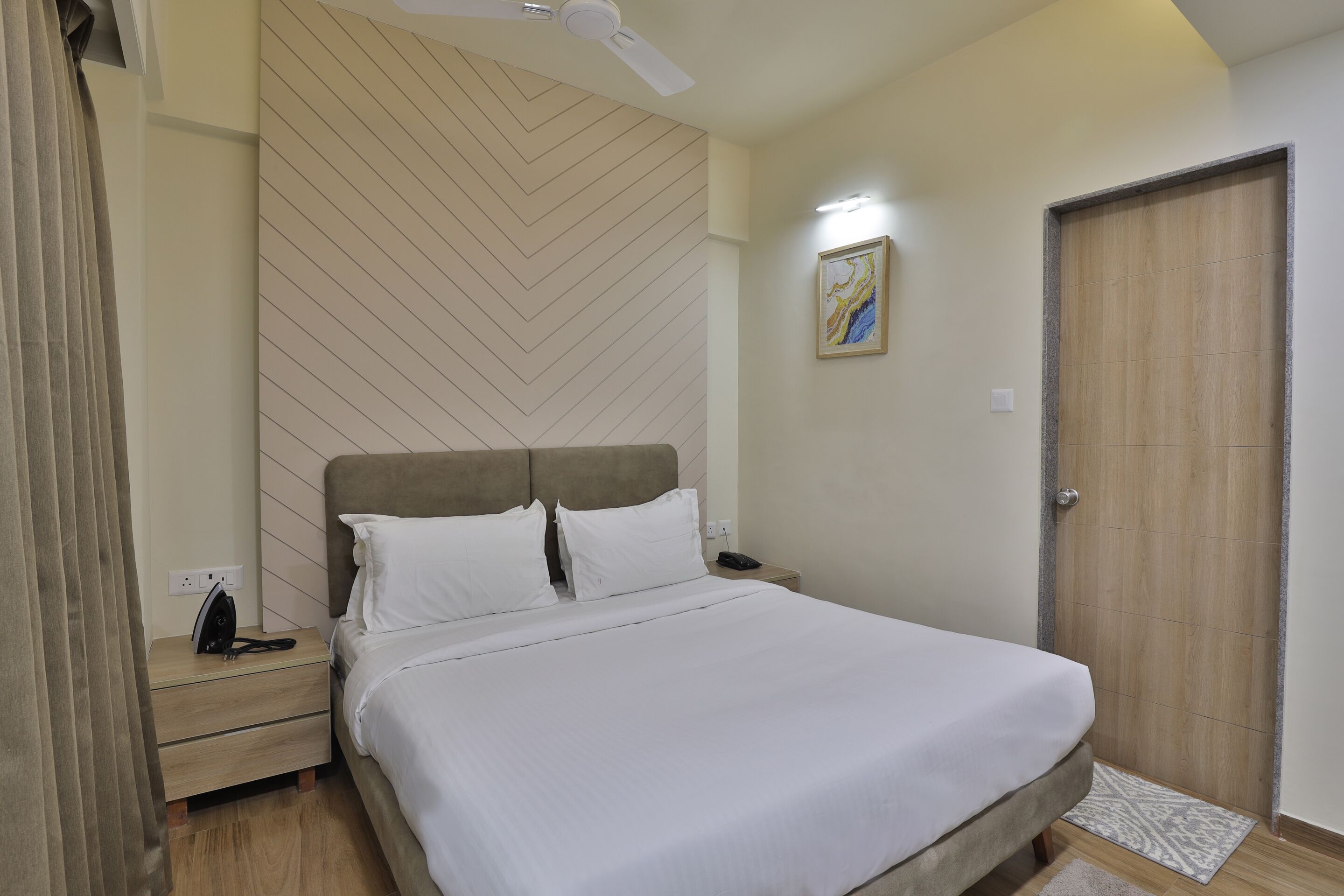 Una cama bien hecha con ropa blanca, un cabecero de madera, una mesita de noche con una lámpara y una puerta con panel de madera.
