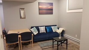 A living room with a blue sofa, a wooden dining table, two chairs, a small black side table, and a painting on the wall. - Pleasant 1 Bedroom Private Suite in the Meadows (Saskatoon)