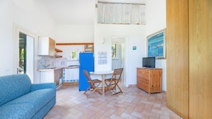 A compact kitchen with a blue refrigerator, a dining table with chairs, a sofa, and a wooden cabinet with a television.