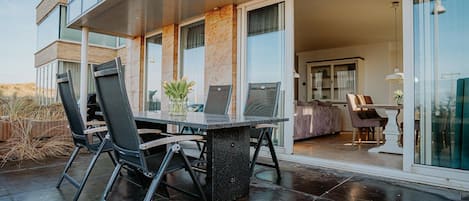 An outdoor dining area with a glass table and chairs, a vase with flowers, and a view of the interior through glass doors.