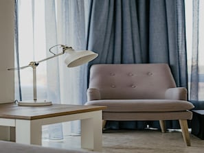 Ein Zimmer mit einem weißen Tisch, einer Lampe, einem Samtstuhl und Blick durch transparente Vorhänge.
