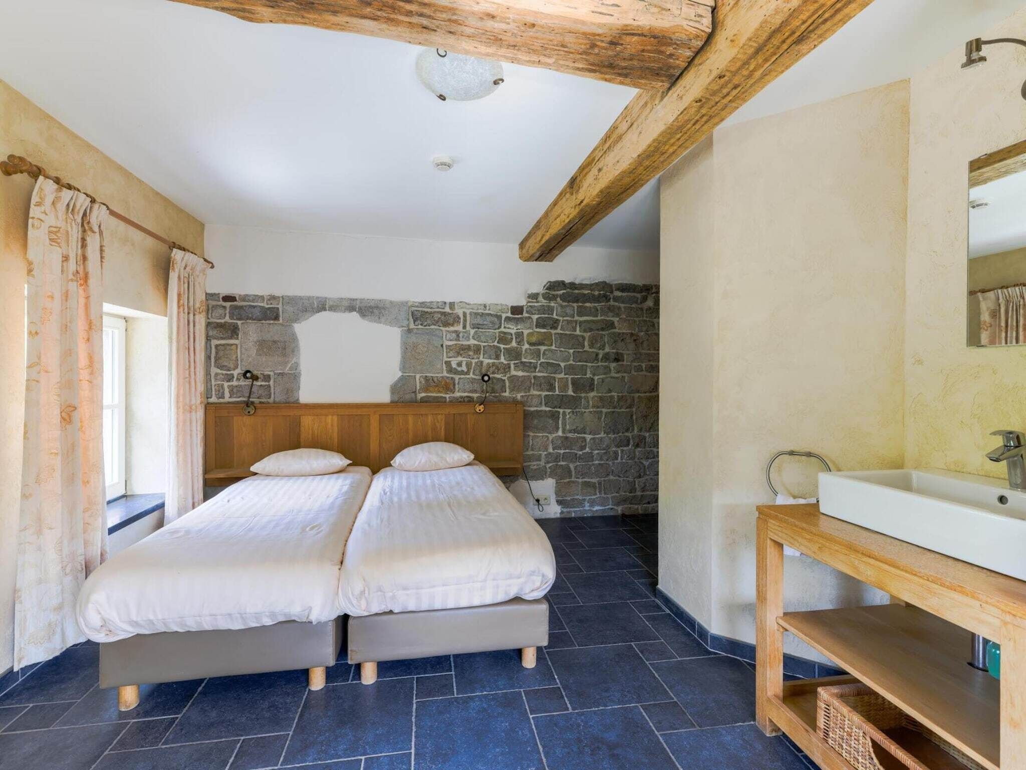 A bedroom with two single beds, a stone wall, a wooden ceiling beam, and a bathroom with a sink and mirror.