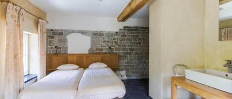 A bedroom with two single beds, a stone wall, a wooden ceiling beam, and a bathroom with a sink and mirror.