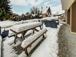 Een buitenruimte bedekt met sneeuw, met houten banken en tafels, een pad van stenen en een gebouw op de achtergrond.