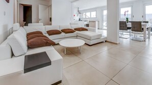 Ein modernes Wohnzimmer mit einem weißen Ecksofa, einem runden Couchtisch und einem Essbereich mit weißem Tisch und Stühlen.