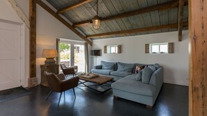 Un salon spacieux avec un grand canapé bleu clair, une table basse en bois et un fauteuil en osier. Le plafond est en bois, avec des poutres apparentes, et on y trouve une lampe ainsi qu’un téléviseur fixé au mur.