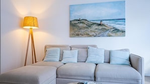 A living room with a sofa, a floor lamp, a painting of a beach, and a coffee table with decorative items. - Apartment in Koudekerke near Seabeach (Dishoek)