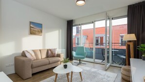 Ein modernes Wohnzimmer mit einer Couch, einem kleinen Tisch und einem Sessel. Ein großes Fenster bietet Blick auf die Gebäude draußen.