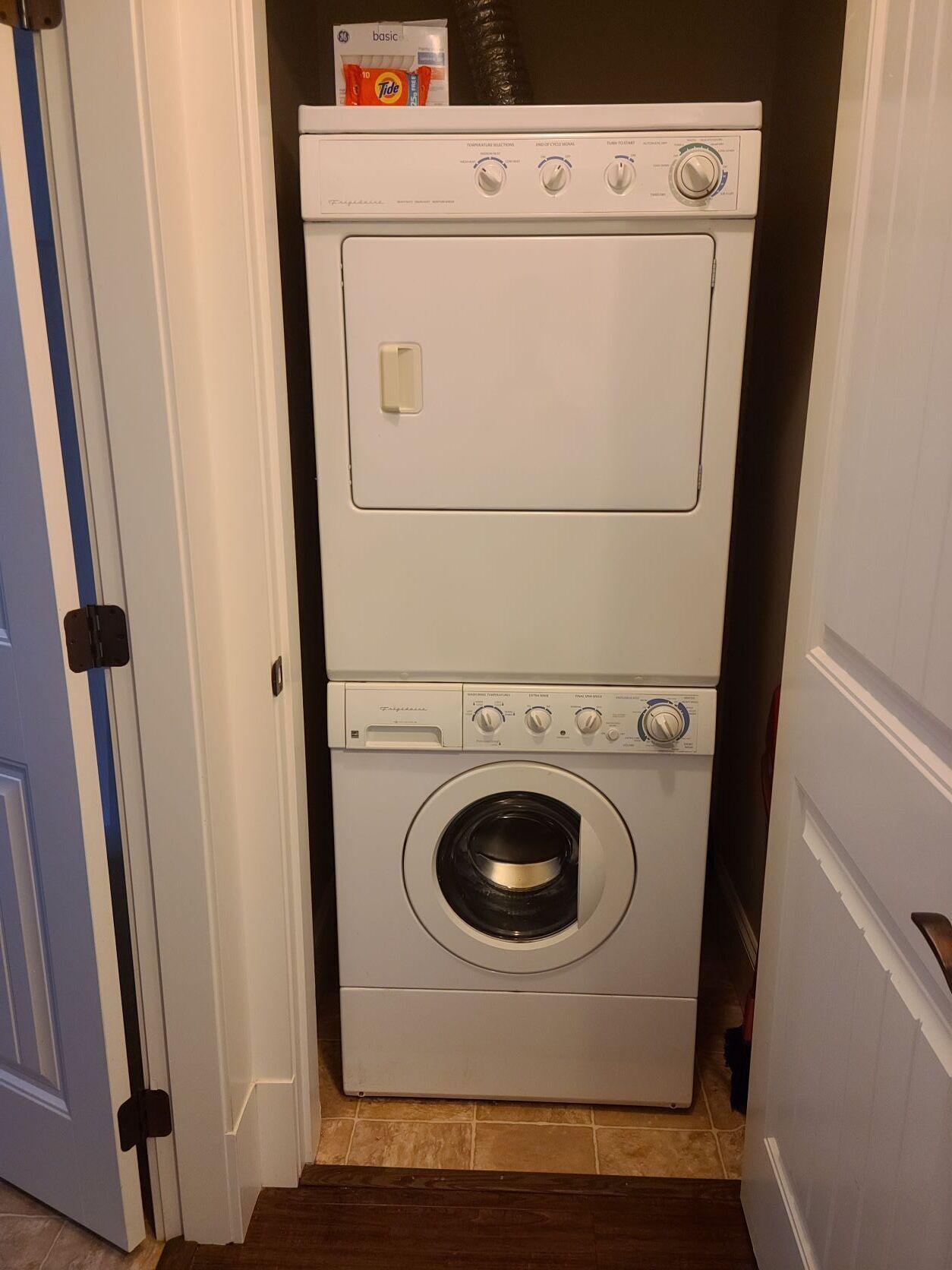 Máquina de lavar e secar frontal compacta, branca e com painel de controle, situada em um corredor estreito com piso de madeira e porta azul.