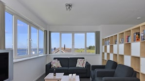 Een moderne woonkamer met een bank, een salontafel en een televisie. Grote ramen bieden uitzicht op zee en een vuurtoren. Een boekenplank staat tegen de muur.