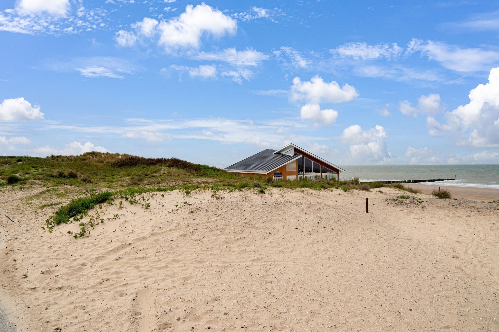 A beach house with a sloped roof, situated on a sandy beach with dunes in the background.