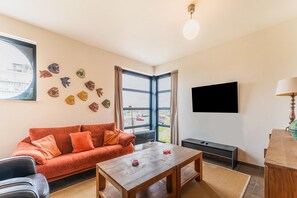 A living room with a red sofa, a wooden coffee table, a TV mounted on the wall, and a window with curtains.