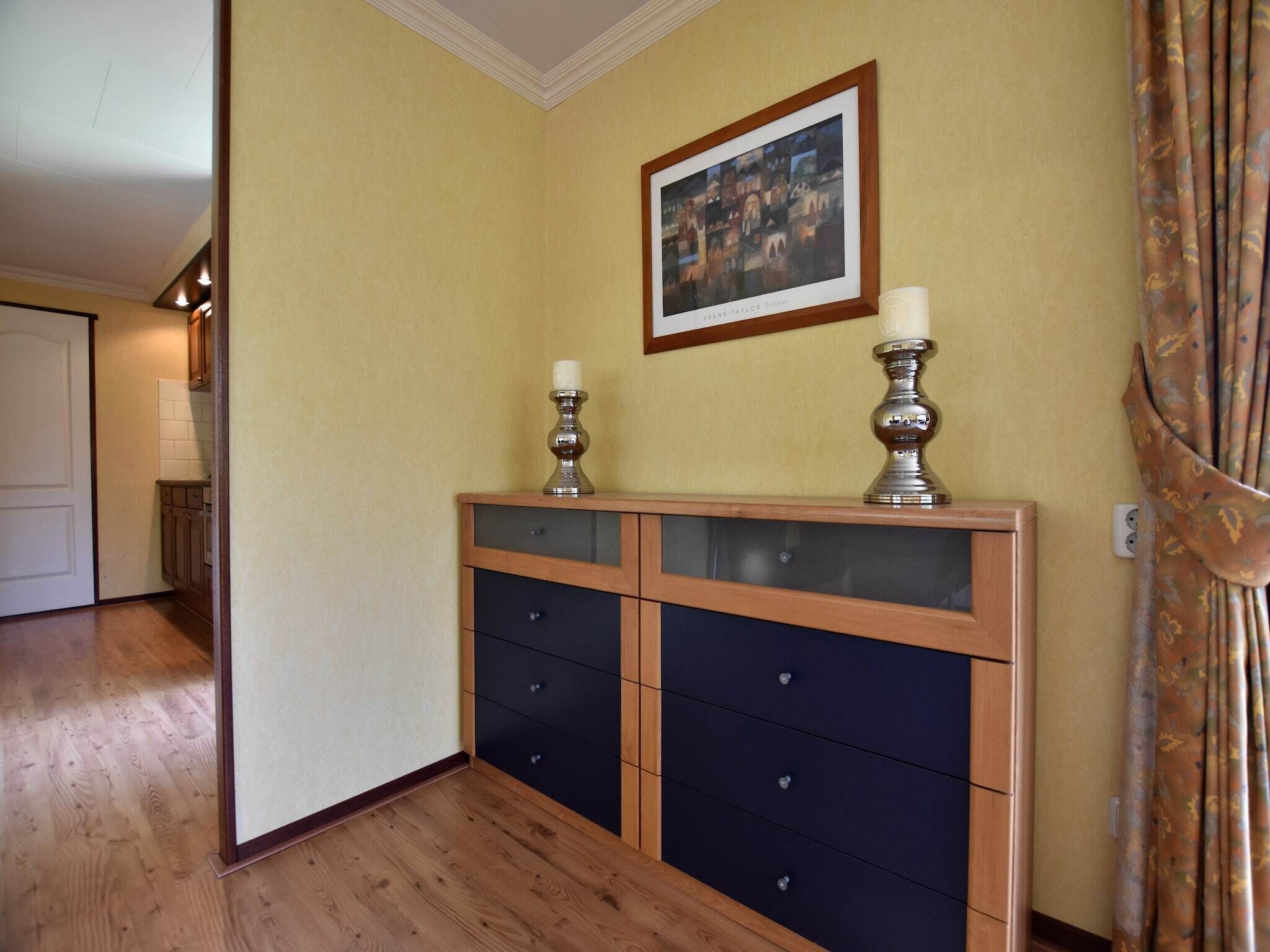 A hallway with a wooden floor, a dark blue cabinet with drawers, a framed artwork on the wall, and a decorative candelabra.