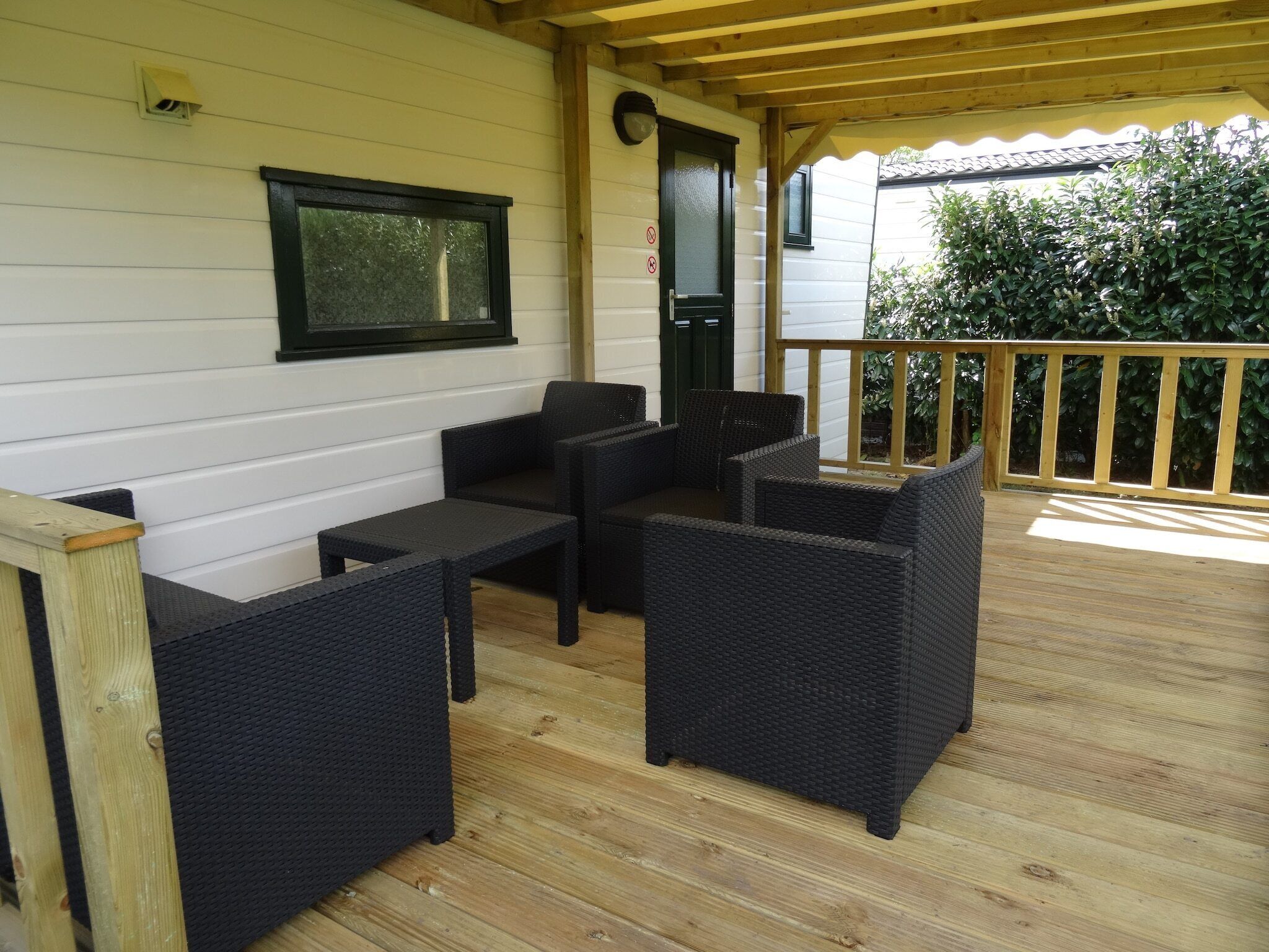 Un área de estar al aire libre cubierta con mobiliario de mimbre, una terraza de madera y un edificio blanco con ventana y puerta de marco negro.
