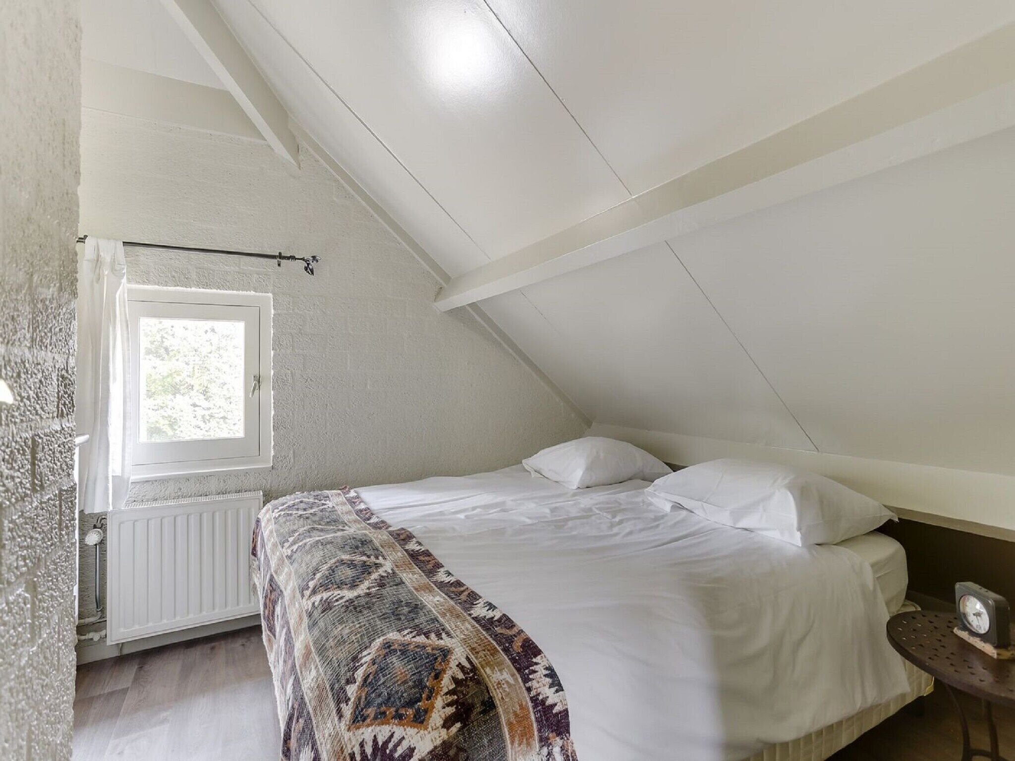 A bedroom with a sloped ceiling, a window, a radiator, a bed with white bedding, and a small table with a clock.