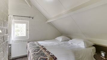 A bedroom with a sloped ceiling, a window, a radiator, a bed with white bedding, and a small table with a clock.
