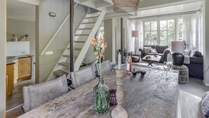 En moderne stue med trappe, et spiseområde med et bord dækket til to og et hyggeligt opholdsområde med sofa og lænestole.