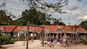 Et sandfyldt udendørs område med trælegekonstruktioner, en bygning med rødt tegltag og en klar himmel.