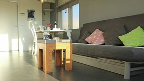 Een moderne woonkamer met een grijze bank, een houten salontafel en een klein keukenhoekje op de achtergrond.