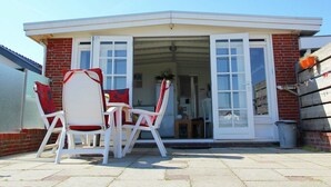 Een terras met witte stoelen en een tafel, een bakstenen gebouw met een witte deur, en een helderblauwe lucht.