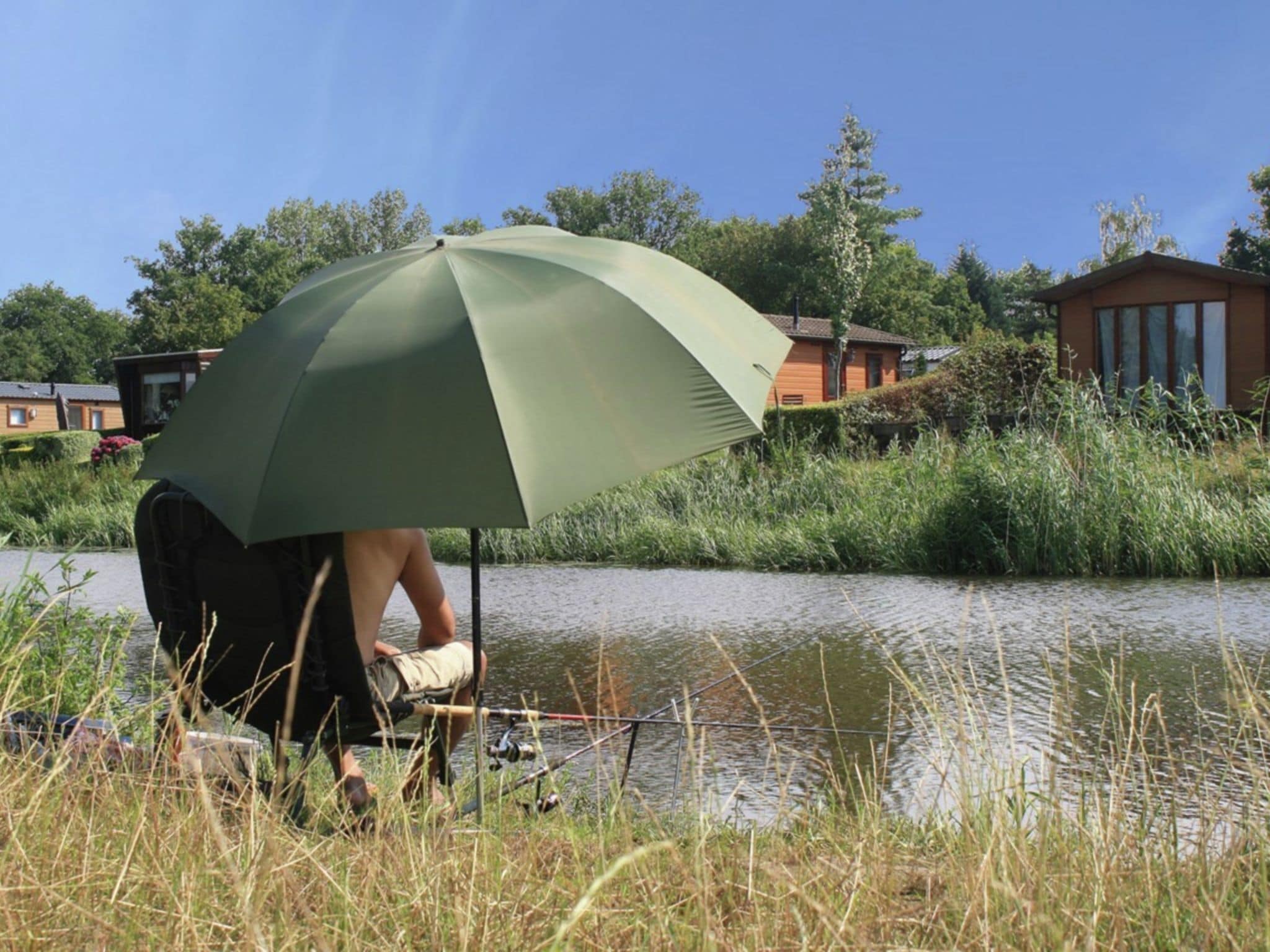 Iemand zit onder een groene parasol aan een rustig waterlichaam, met huizen op de achtergrond.