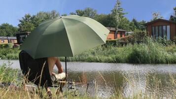 Una persona sentada bajo un paraguas verde junto a un cuerpo de agua tranquilo, con casas al fondo.