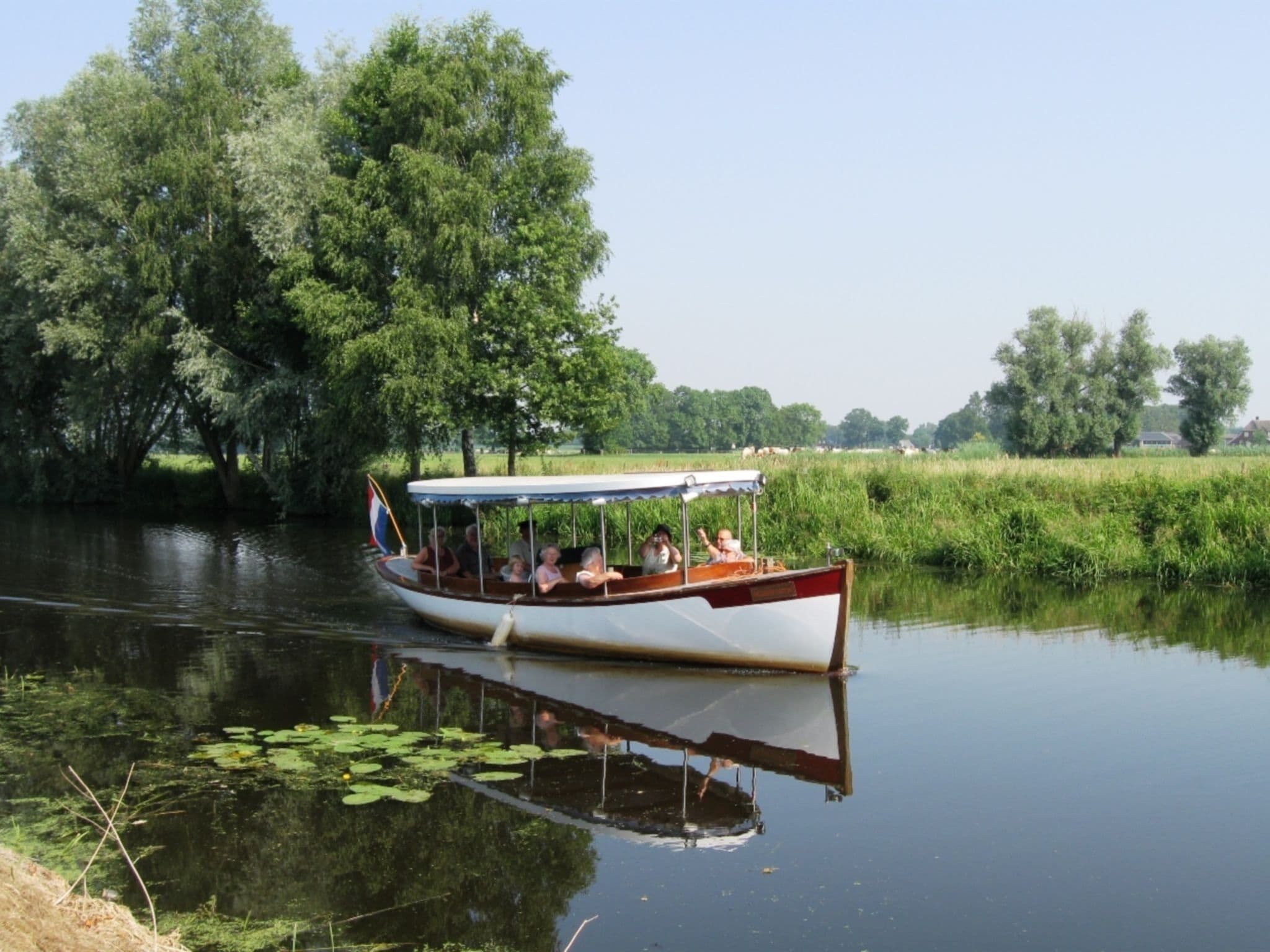 A narrowboat on a calm river with passengers, surrounded by lush greenery and trees.