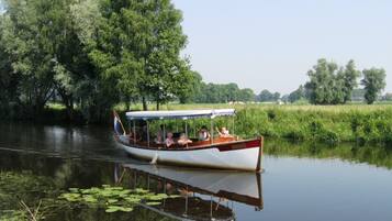 A narrowboat on a calm river with passengers, surrounded by lush greenery and trees.