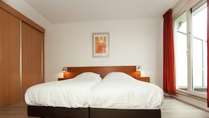 A double bed with a wooden headboard, two bedside tables with lamps, a large window with red curtains, and a wooden wardrobe.