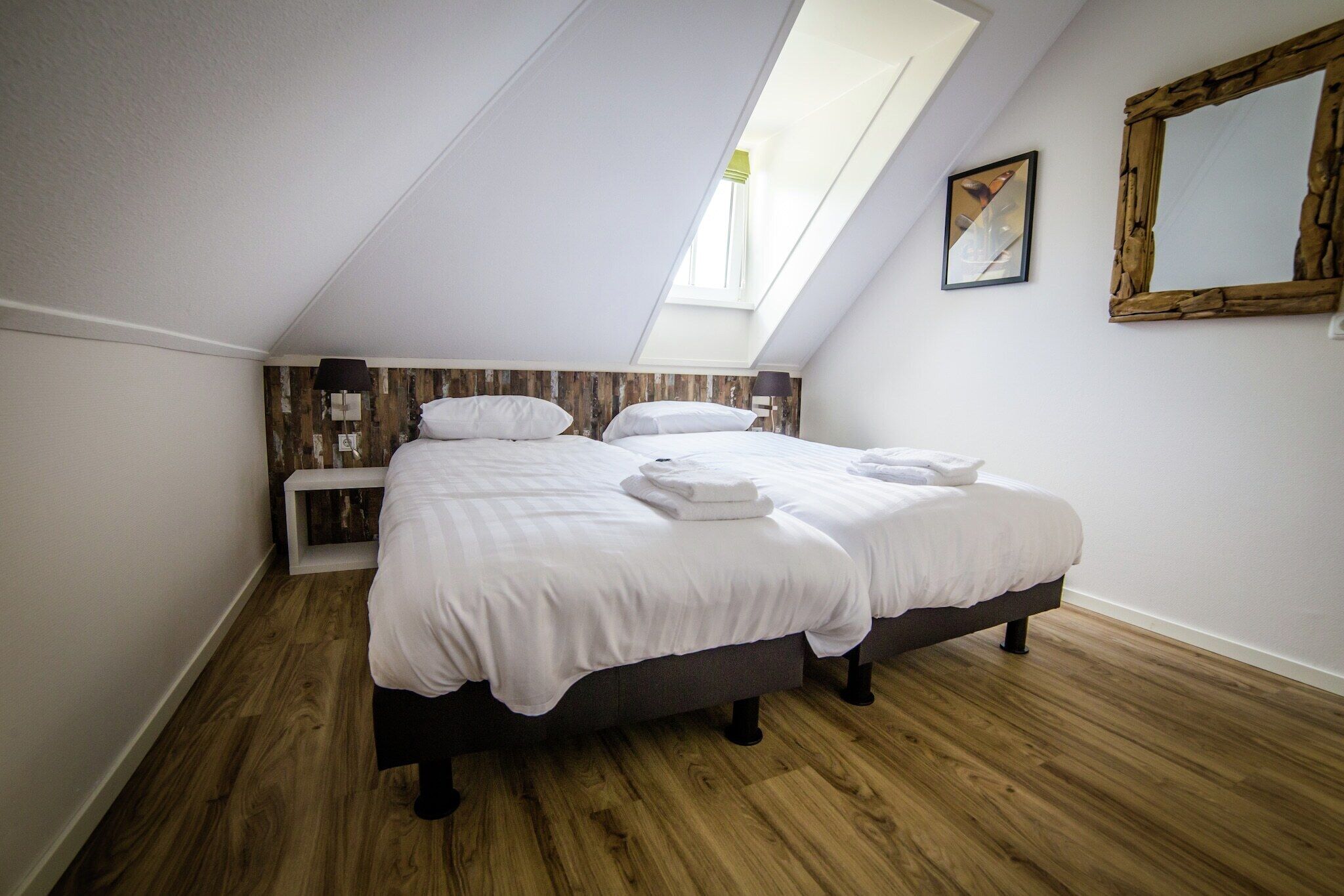 A bedroom with a skylight, a bed with white linens, a bedside table, and a mirror on the wall.