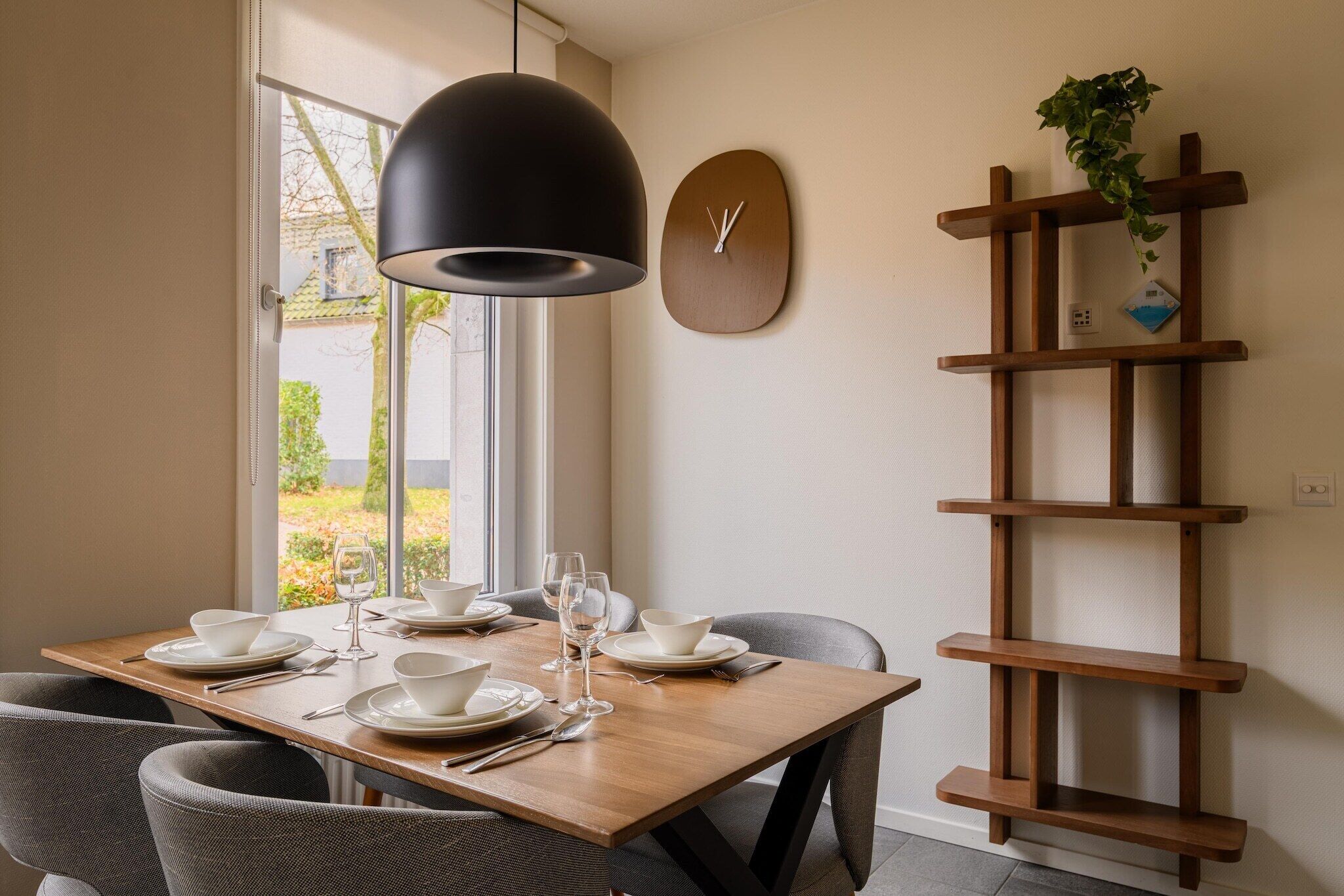 Comedor con mesa de madera dispuesta para cuatro personas, una lámpara colgante negra, un reloj de pared y una estantería de madera con una planta.