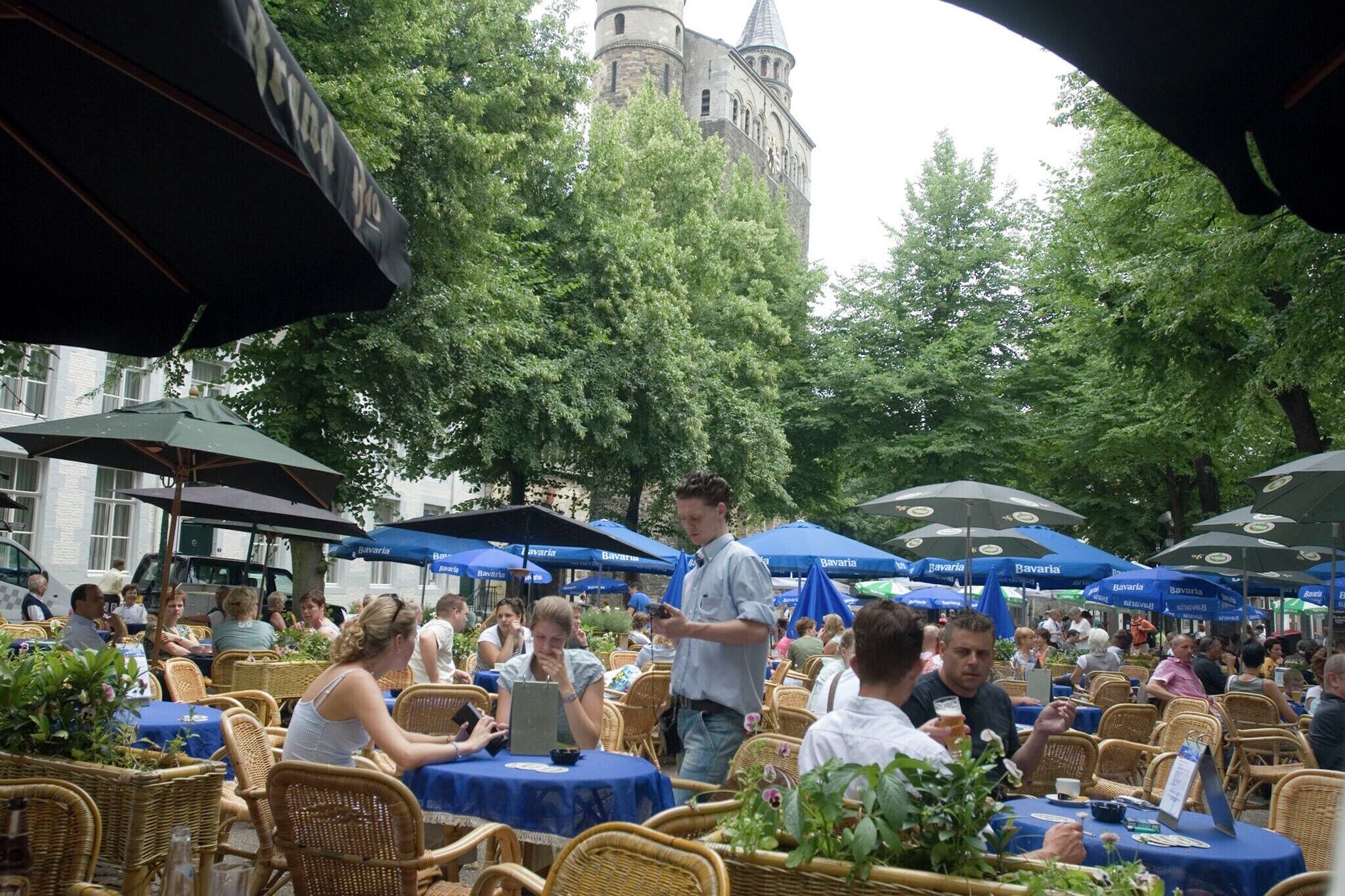 Ein Essbereich im Freien mit Korbstühlen und blauen Sonnenschirmen, umgeben von Bäumen und einem historischen Gebäude im Hintergrund.