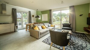 A modern living room with a sofa, armchair, and dining table. There is a kitchen area with cabinets and a stove. The room has large windows with curtains, and a view of greenery outside. - Stylish Villa in Maastricht (Maastricht)