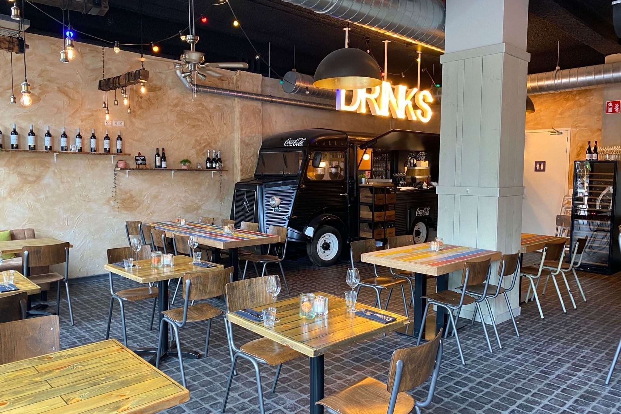 Een restaurantinterieur met houten tafels en stoelen, een bar met flessen en een oude Coca-Cola vrachtwagen.