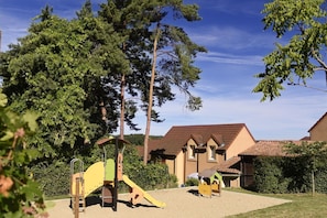 Une aire de jeux avec un toboggan et des structures d’escalade, entourée d’arbres et bordée d’un bâtiment doté d’un toit en tuiles.